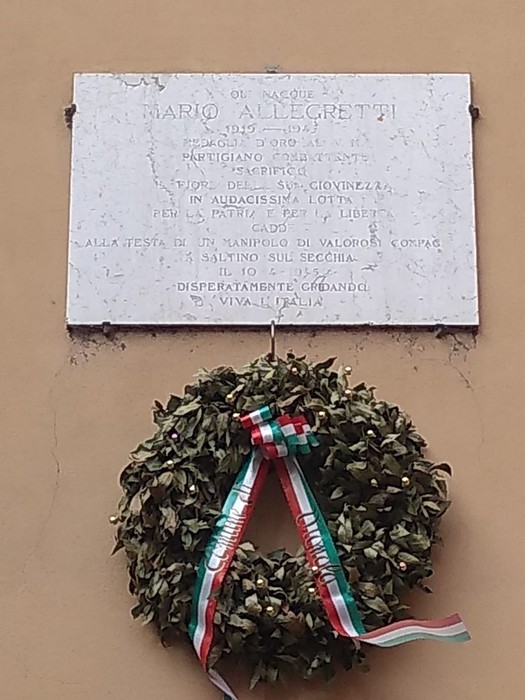 Lapide commemorativa di Mario Allegretti con un'omaggio floreale e un nastro tricolore.