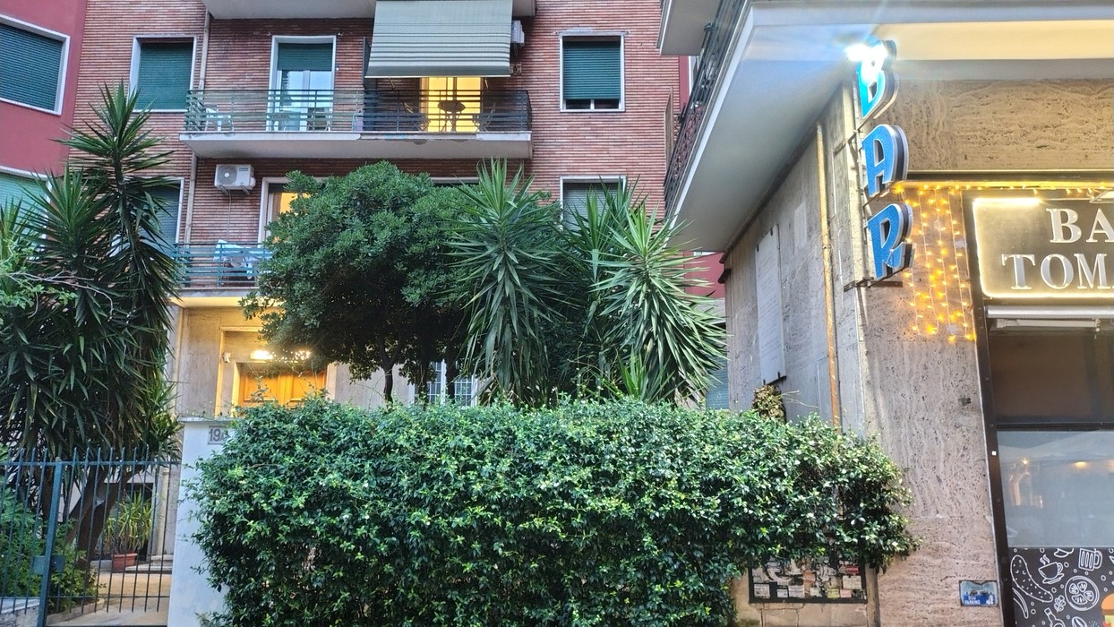 Edificio residenziale con piante verdi e un bar con insegna luminosa, situato in un contesto urbano.