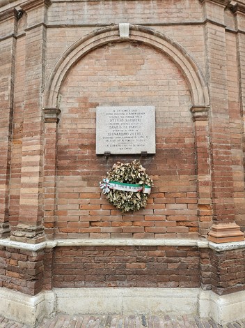 Lapide commemorativa dedicata a Barbieri, De Marchi e Zelioli, con una corona di fiori sotto una struttura in mattoni.