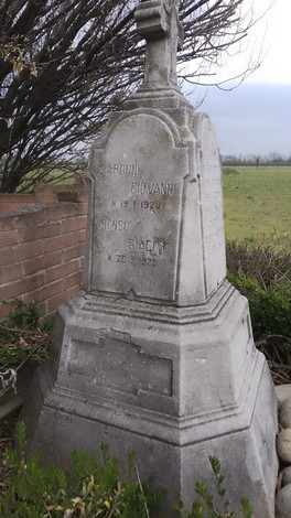 Cippo commemorativo dedicato ai partigiani di Camuzzago, tra vegetazione e un campo aperto sullo sfondo.