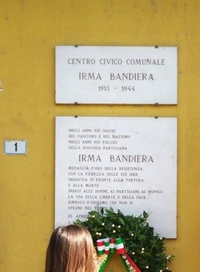 Lapide commemorativa dedicata a Irma Bandiera, con un omaggio floreale e un muro di fondo giallo.
