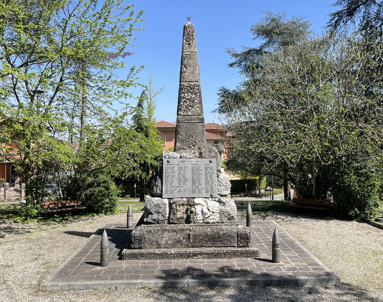 Monumento a obelisco in pietra con lapide incisa, su basamento, circondato da alberi in un parco.