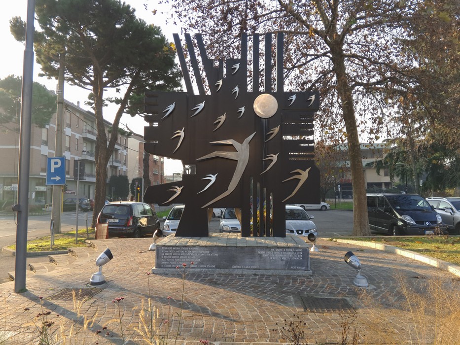 Monumento in metallo scuro con sagome di uccelli stilizzati, su basamento con iscrizione, in una piazza alberata.