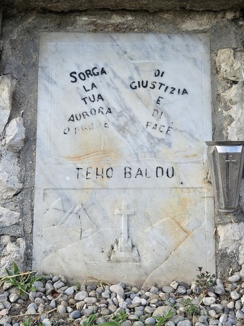 Cippo commemorativo con iscrizioni su una parete di pietra, circondato da ciottoli e vegetazione.