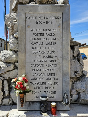 Monumento ai Caduti con nomi incisi, circondato da rocce e decorato con fiori e candele.