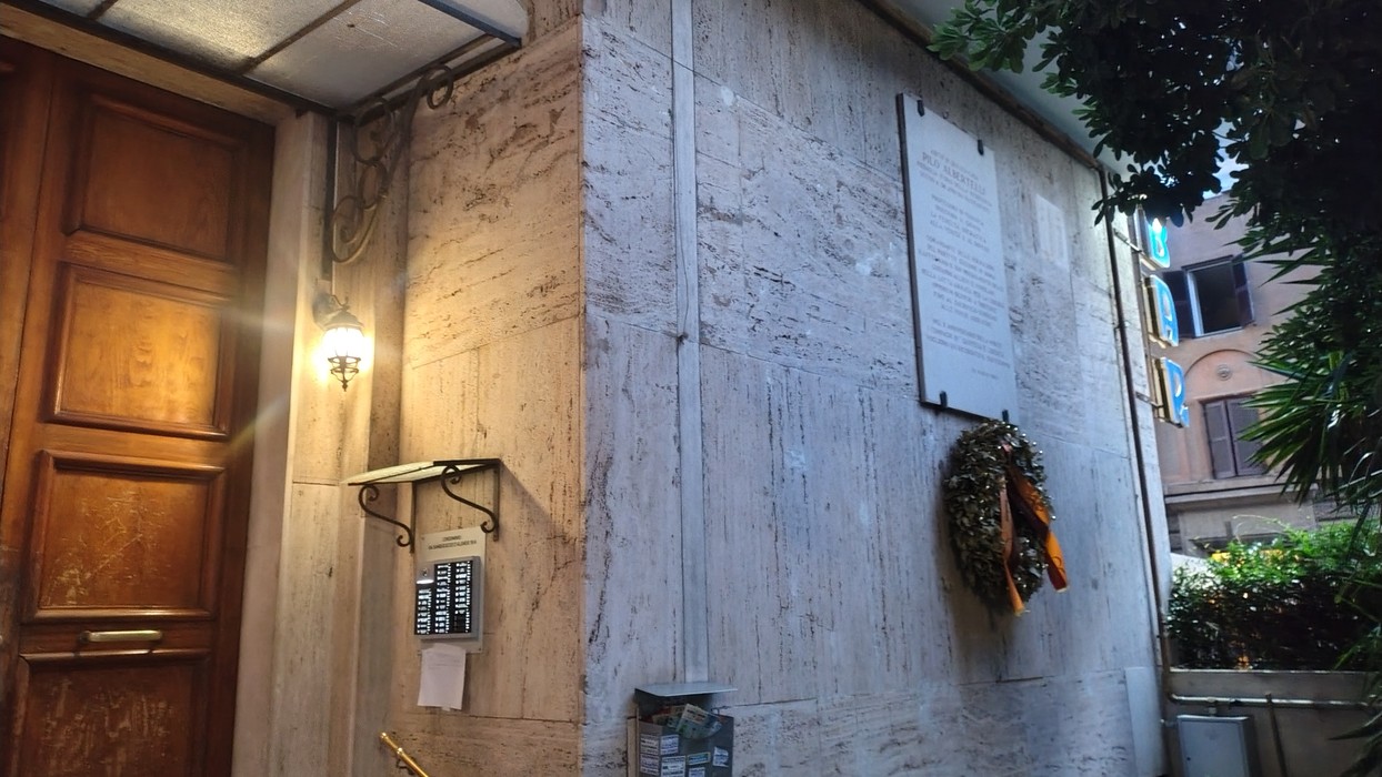 Lapide commemorativa su una parete, con una corona di fiori e un ingresso in legno sullo sfondo.