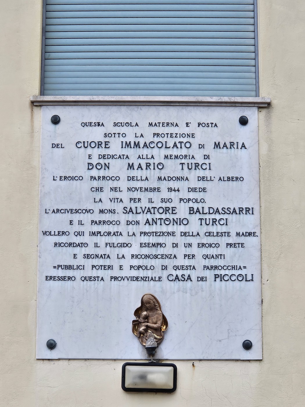 Lapide commemorativa dedicata a Don Mario Turci, con riferimento alla protezione di Maria e alla vita della comunità.