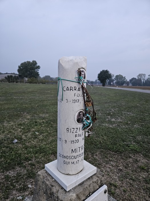 Cippo cilindrico in pietra con iscrizioni e fotografie dei caduti, posto su un prato in aperta campagna.
