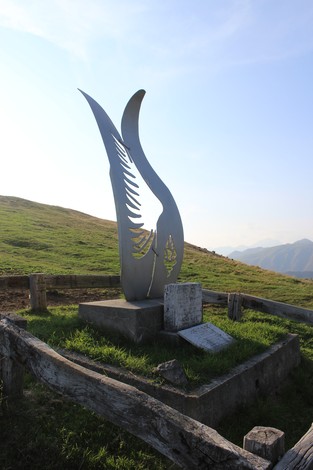 Monumento all'aviolancio di Cadorna, scultura stilizzata su un prato verde, sullo sfondo colline e cielo sereno.