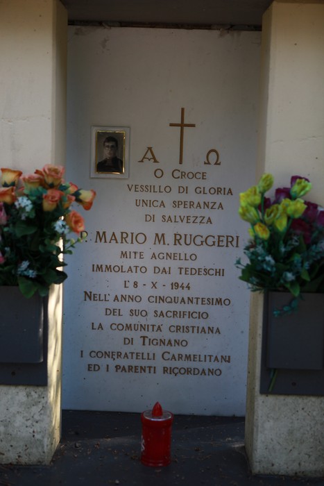 Cippo commemorativo dedicato a Mario M. Ruggeri, con fiori e una croce, in ricordo dei caduti della Seconda Guerra Mondiale.