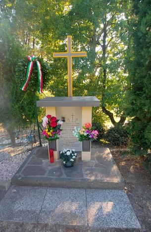 Cippo commemorativo con fiori e una croce, immerso nella vegetazione. Un tributo alla memoria.