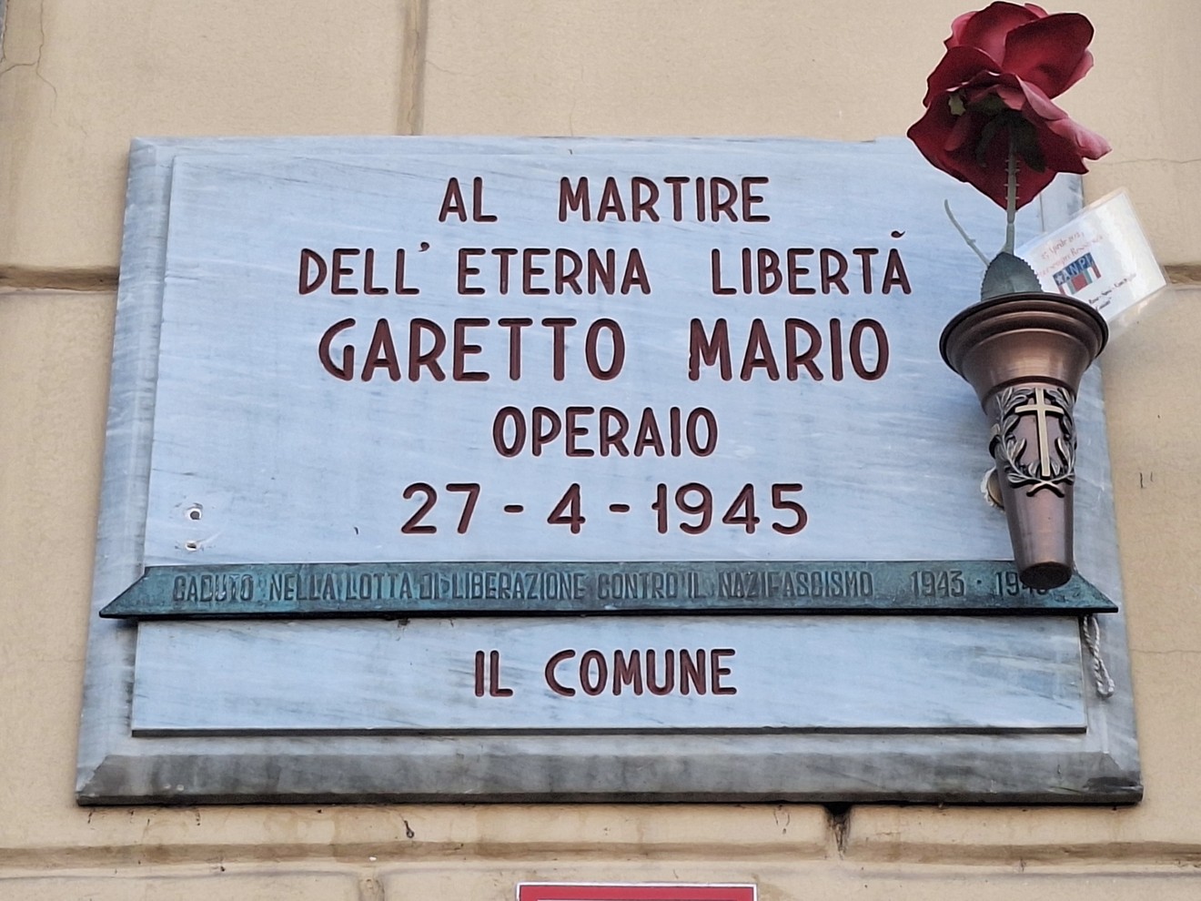 Lapide marmorea su una parete, con iscrizione e una fiaccola metallica sormontata da un fiore rosso.