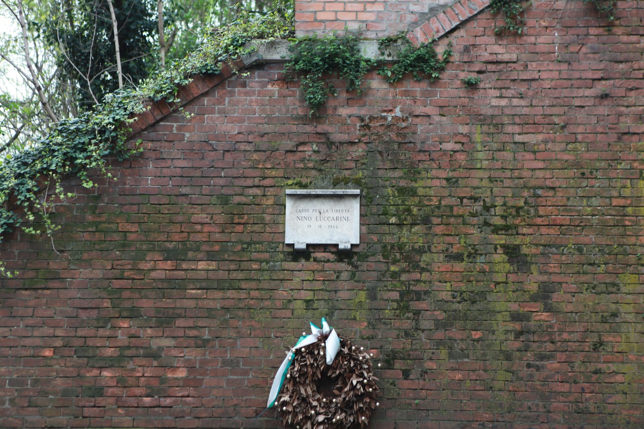 Lapide commemorativa su un muro di mattoni, decorata con una corona di foglie e un nastro.