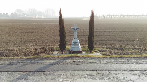 Croce commemorativa con cipressi, circondata da un vasto campo arato e un paesaggio rurale.