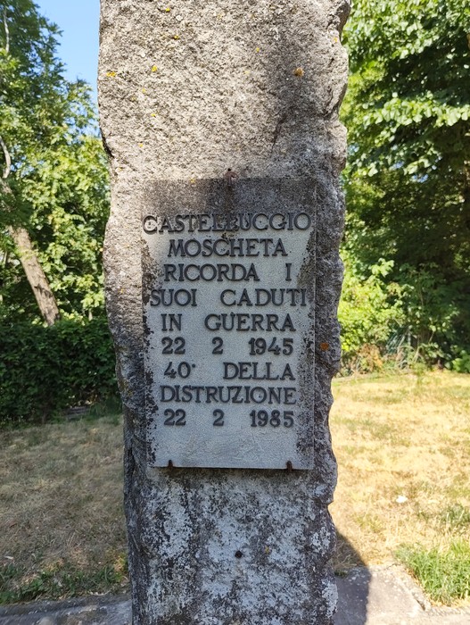 Targa in marmo con iscrizione fissata su un pilastro di pietra, circondata da vegetazione.