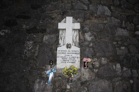 Lapide commemorativa di Carmelo Baronchelli, con fiori e una croce, situata su una parete di pietra.