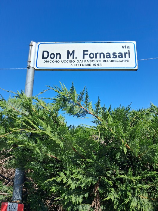 Cippo commemorativo dedicato a Don M. Fornasari, con sfondo di cielo blu.