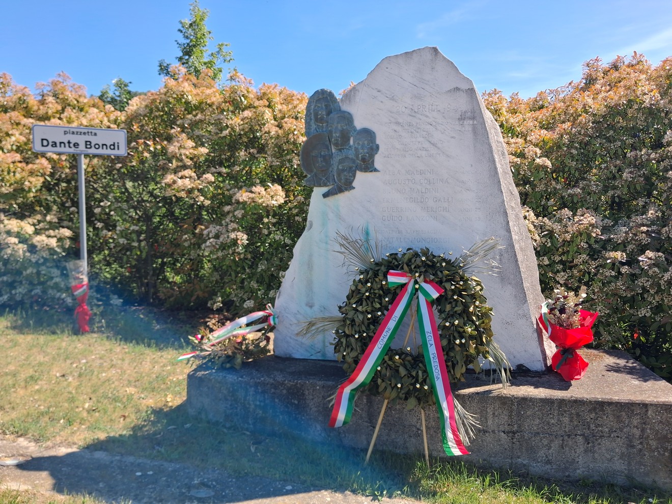 Monumento per la Liberazione con corona d'alloro, circondato da fiori e cartelli, in una giornata di sole.