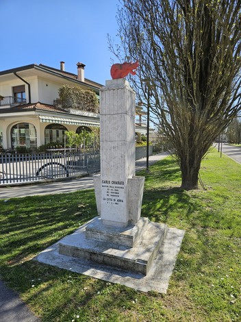 Monumento a Chiarato con scultura rossa su base di marmo, immerso nel verde e vicino a una residenza.