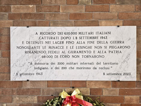 Lapide in marmo su parete di mattoni, con iscrizione e corona di fiori appoggiata sotto.