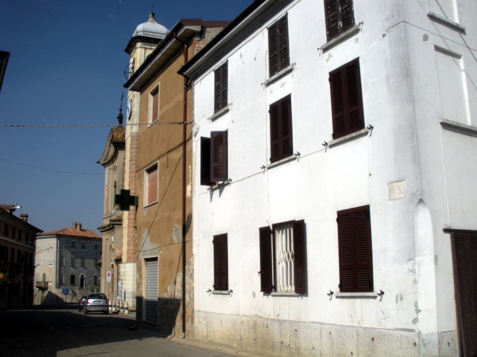 Lapide commemorativa di Fugazza Mario in una via storica, con edifici caratteristici e cielo sereno.