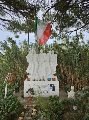 Monumento ai Martiri della Macchinina, con tre figure scolpite e decorazioni floreali, circondato da vegetazione.
