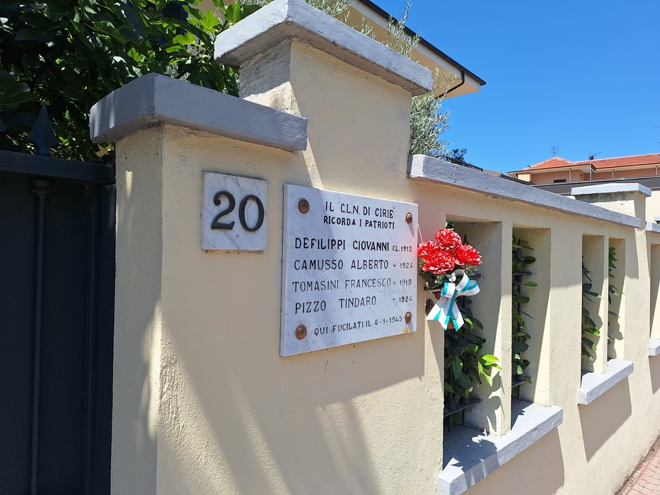 Lapide con iscrizione su pilastro di un muro di cinta, con fiori artificiali appesi accanto.