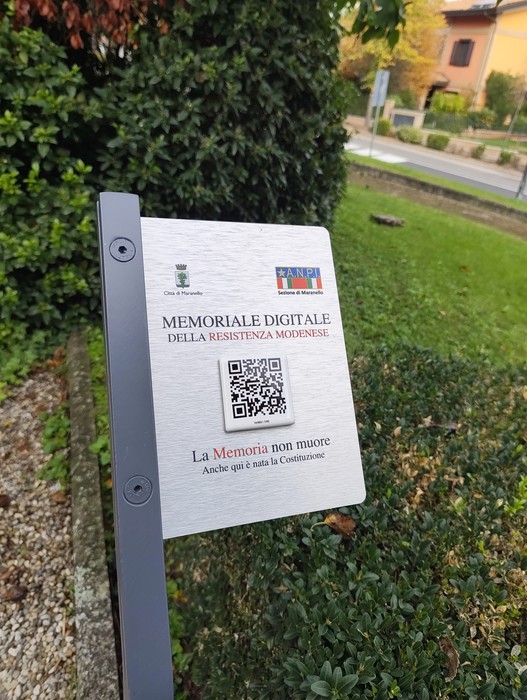 Targa metallica con QR code su palo, davanti a una siepe lungo una strada alberata.