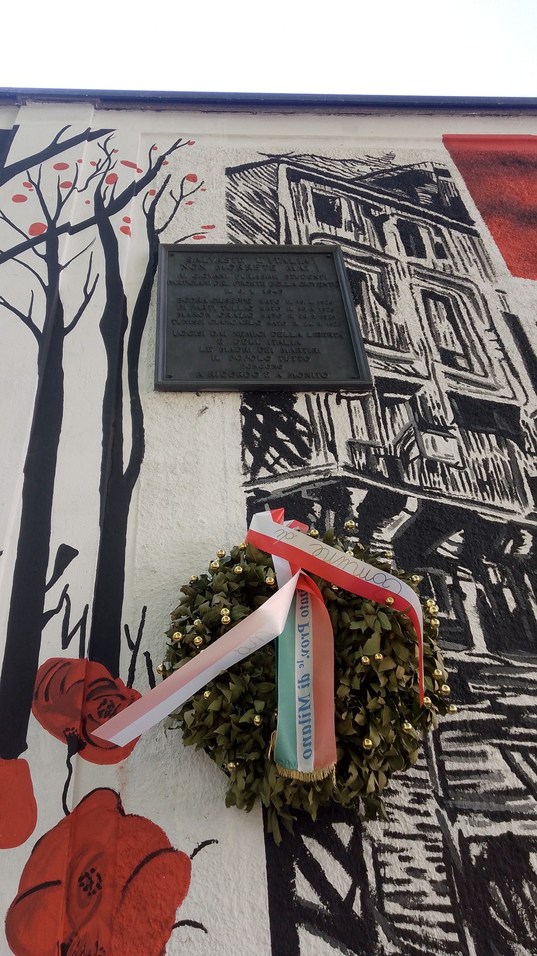 Lapide commemorativa con decorazione floreale, affissa su un muro decorato da murales, dedicata a eventi storici.