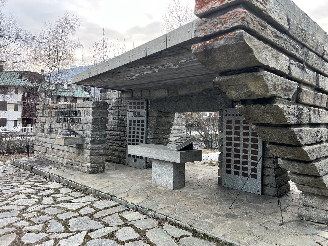 Monumento in pietra con copertura a sbalzo, pannelli forati e lapide con iscrizione, su piazza lastricata.