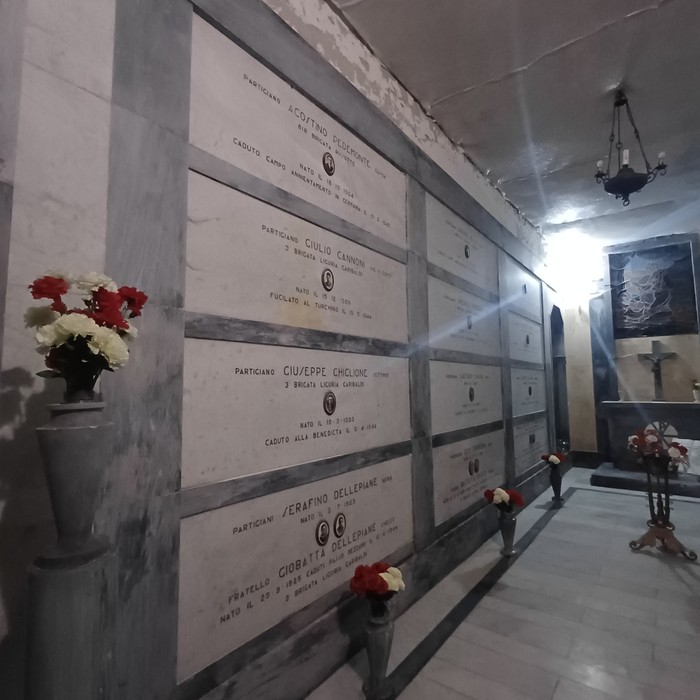 Parete interna con lapidi in marmo e iscrizioni, fiori deposti su mensole e vasi lungo il corridoio.