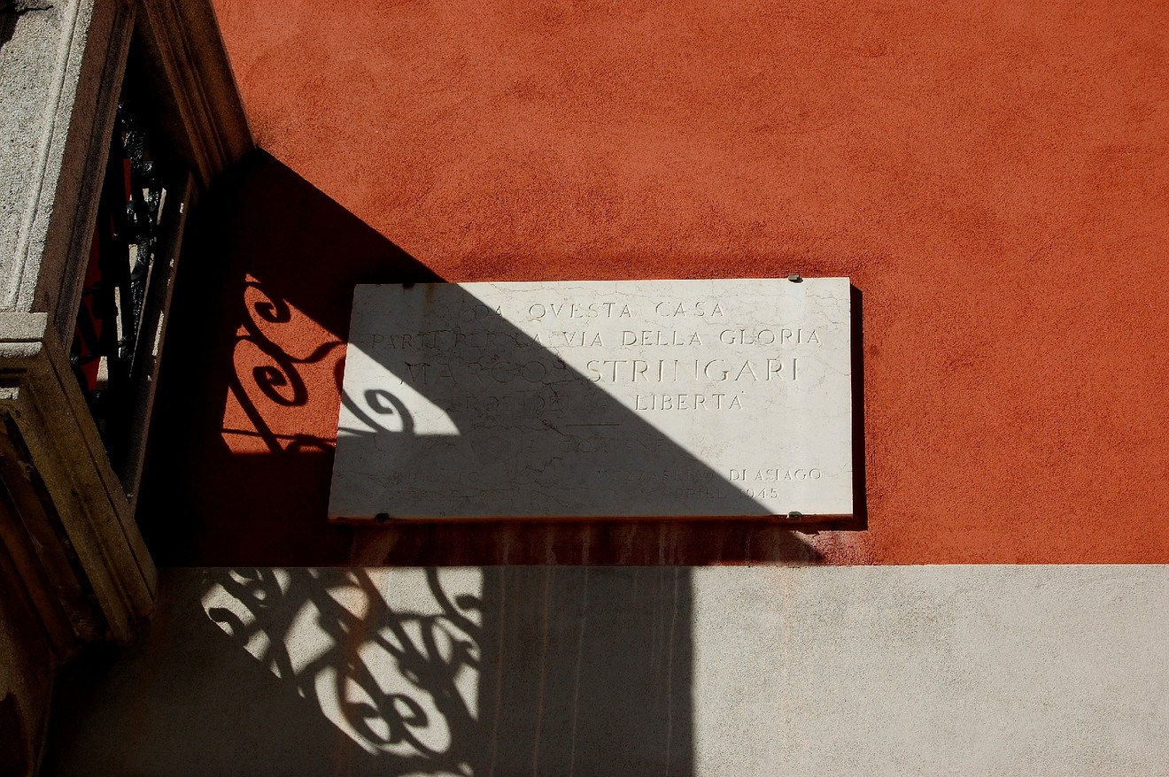Lapide commemorativa a Stringari su parete arancione, con ombre e dettagli architettonici in evidenza.
