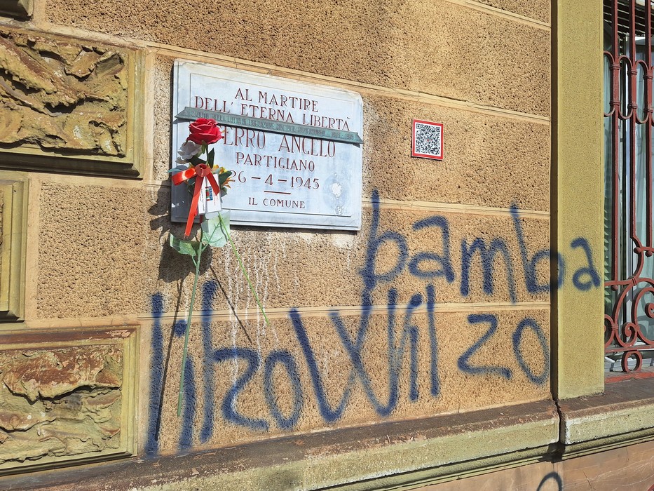 Lapide in marmo su parete, con iscrizione, fiori deposti sotto e scritte murali sovrapposte.