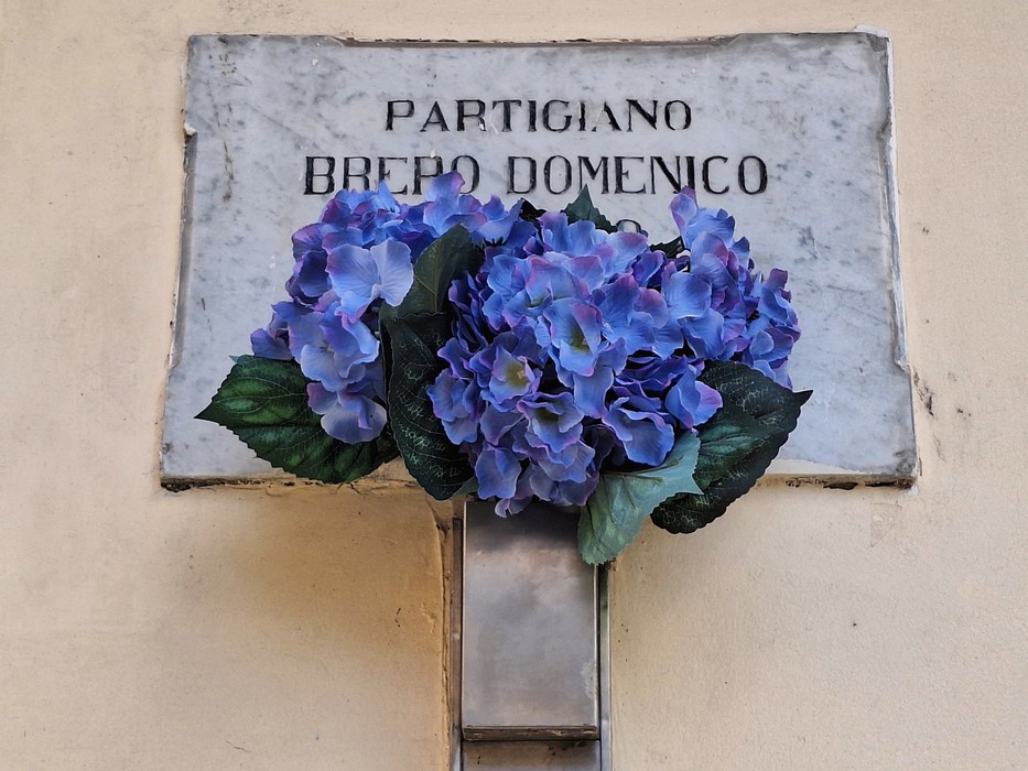 Lapide commemorativa a Brero con fiori blu artificiali, posizionata su una parete.
