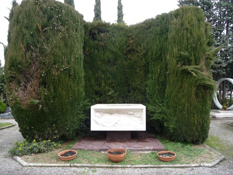 Monumento funebre a Zambianchi, composto da una lastra bianca e circondato da alti arbusti verdi.