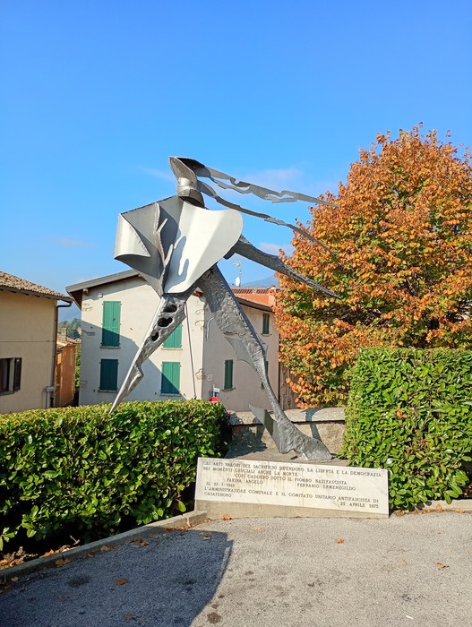 Monumento in acciaio dalle forme astratte e slanciate, su siepe squadrata, con lapide e iscrizione alla base.