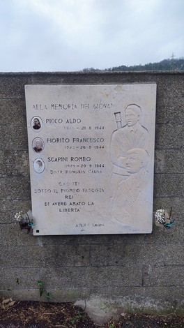 Lapide commemorativa con nomi e date di giovani caduti per la libertà. Sfondo grigio e atmosfera serena.