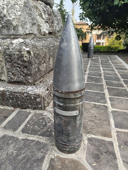Proiettile di artiglieria in metallo eretto su pavimentazione in pietra, davanti a un basamento roccioso.