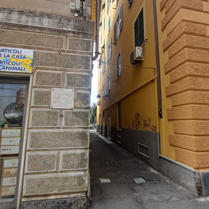 Lapide con iscrizione su parete in pietra, all'imbocco di un vicolo stretto tra edifici.