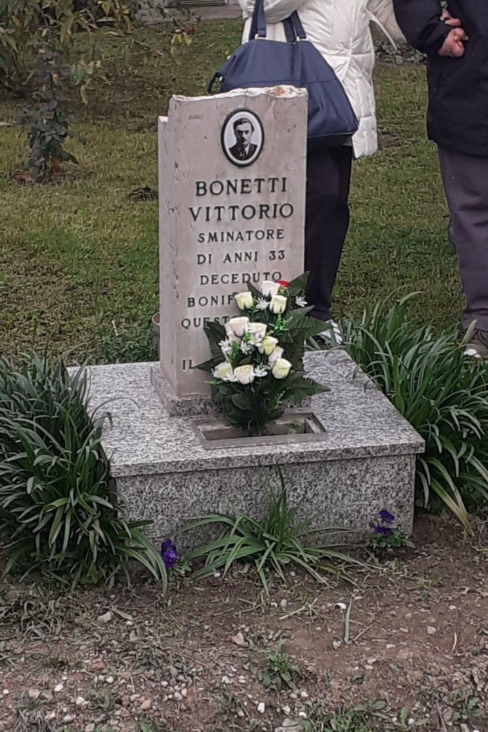 Cippo commemorativo di Vittorio Bonetti, con fiori freschi e persone che si raccolgono in ricordo.