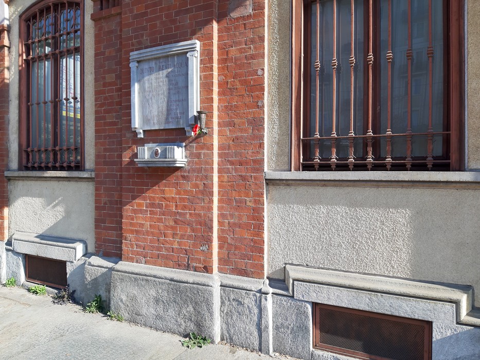Lapide storica affissa a un muro di mattoni rossi, con finestre decorate in ferro.