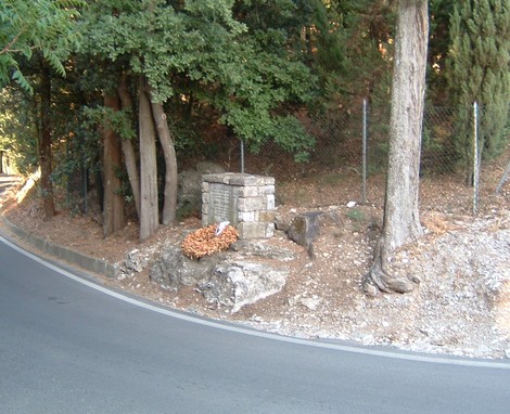 Cippo commemorativo immerso nel verde di una strada, circondato da alberi e natura.