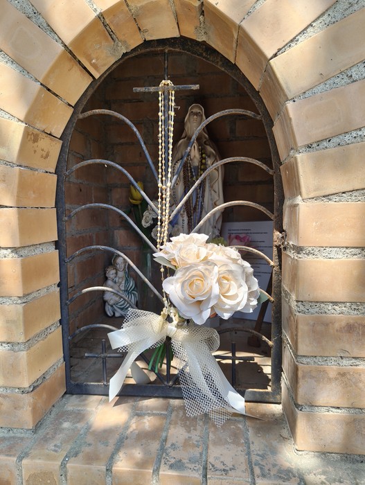 Nicchia ad arco in mattoni con statua della Madonna, rosario e bouquet di rose bianche appesi alla grata.