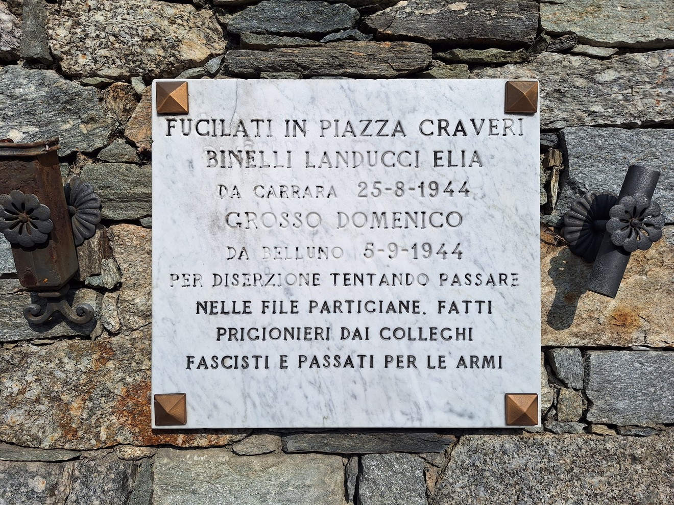 Lapide marmorea con iscrizione, fissata a un muro in pietra, affiancata da due elementi in ferro battuto.