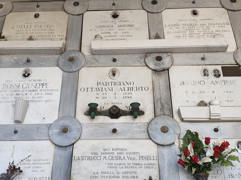 Parete cimiteriale con lapidi marmoree di partigiani, ciascuna con iscrizione e fiori davanti ad alcune tombe.