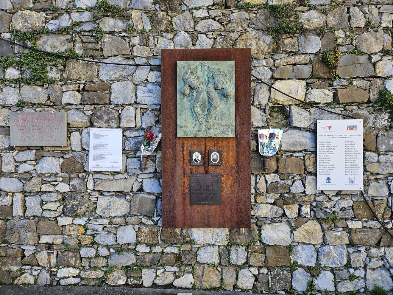 Targhe informativa e artistica su un muro di pietra a Piazza del Popolo, con elementi decorativi e varie iscrizioni.