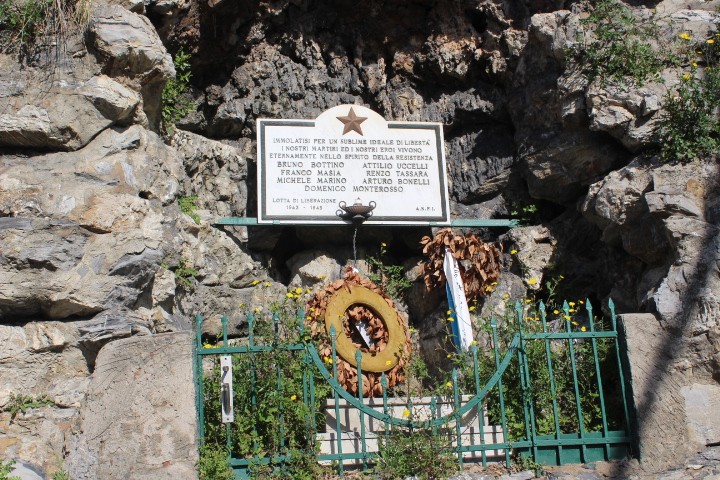 Lapide commemorativa ai caduti per la Libertà, incastonata tra le rocce, con una corona d'alloro e decorazioni floreali.