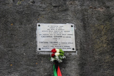Lapide commemorativa dedicata a Cazzaniga e Fontana, con decorazioni floreali, su una parete di pietra.