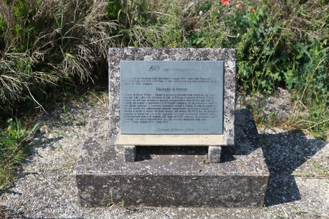 Croce delle Pradole, monumento in pietra circondato da vegetazione, con pannello esplicativo su base di cemento.