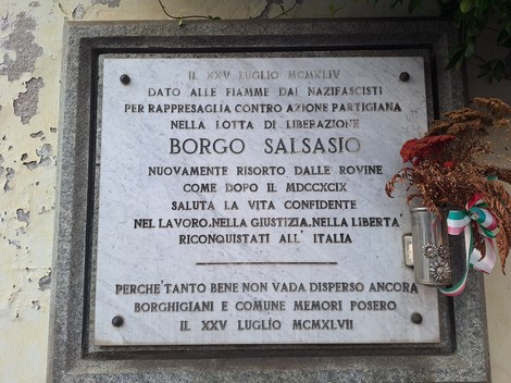 Lapide in marmo con iscrizione, fissata a una parete esterna, con fiori secchi accanto